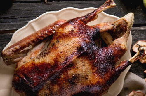 Roast stuffed goose on ceramic plate with ripe apples over wooden kitchen table. Dark rustic style. Top view.